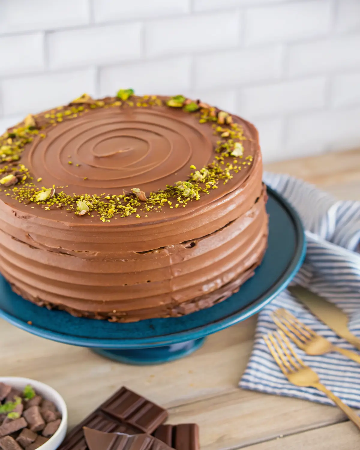 dubai-chocolate-cake-whole-pistachio-frosting Dubai Chocolate Cake with smooth chocolate frosting and pistachio topping on a blue stand